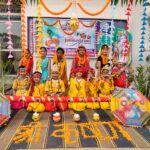 Children in yellow and orange costumes pose for janmashtami at school celebration with marigold rangoli and bright umbrellas in the background